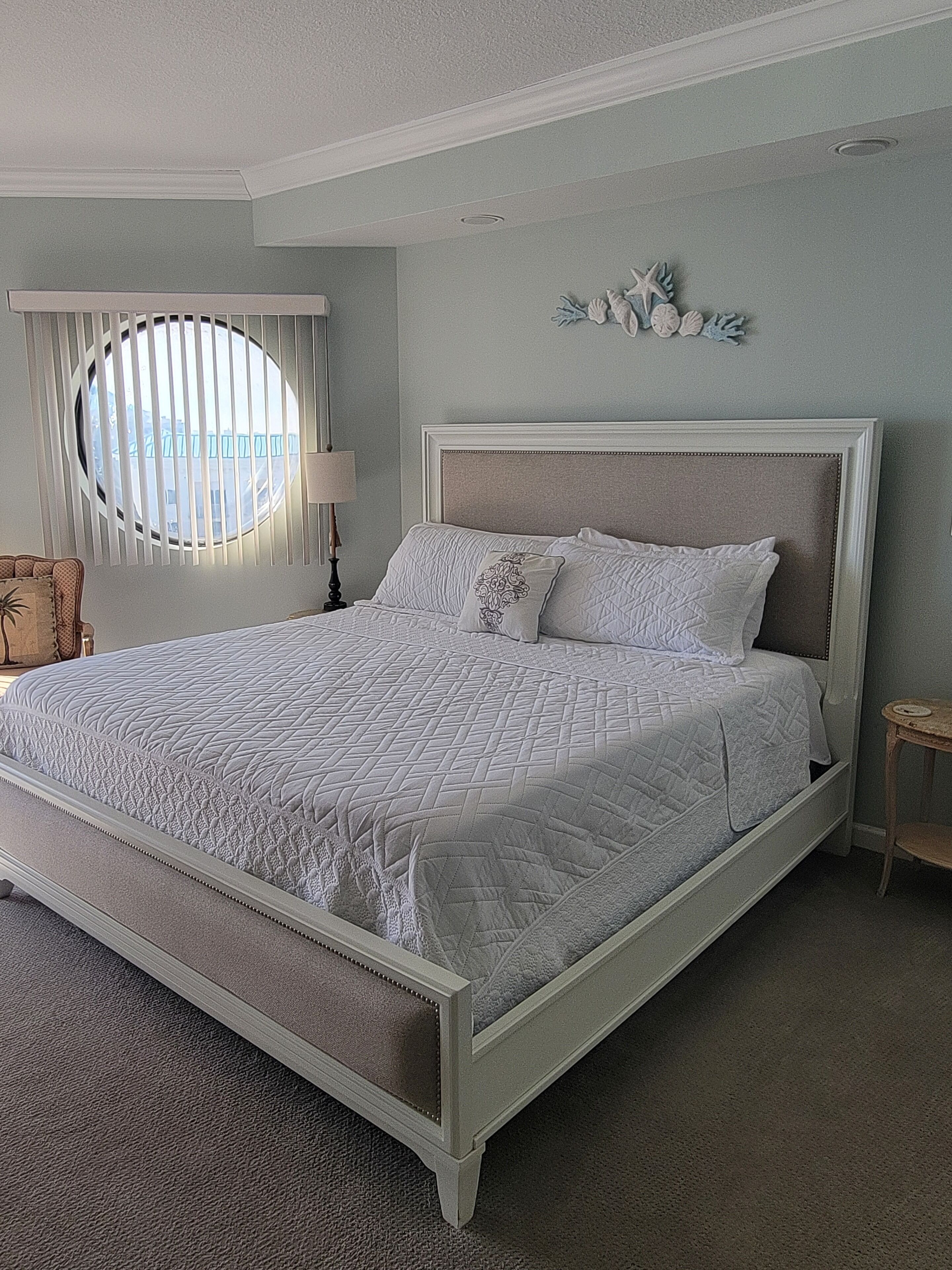 A bedroom with a large bed, a round window, a nightstand, and a chair.