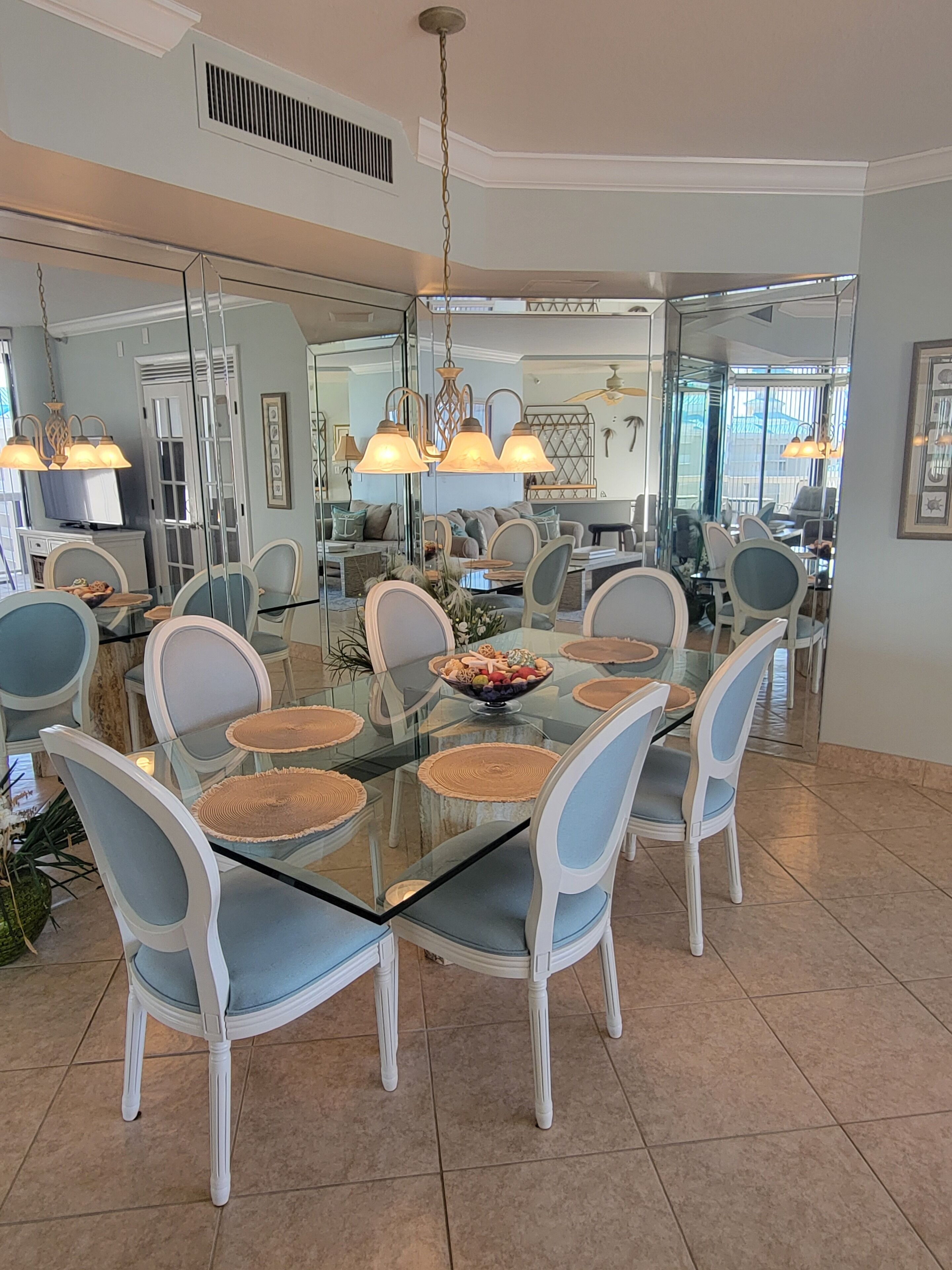 Un coin repas avec une table en verre entourée de chaises blanches, un lustre central et un saladier de fruits posé sur la table.