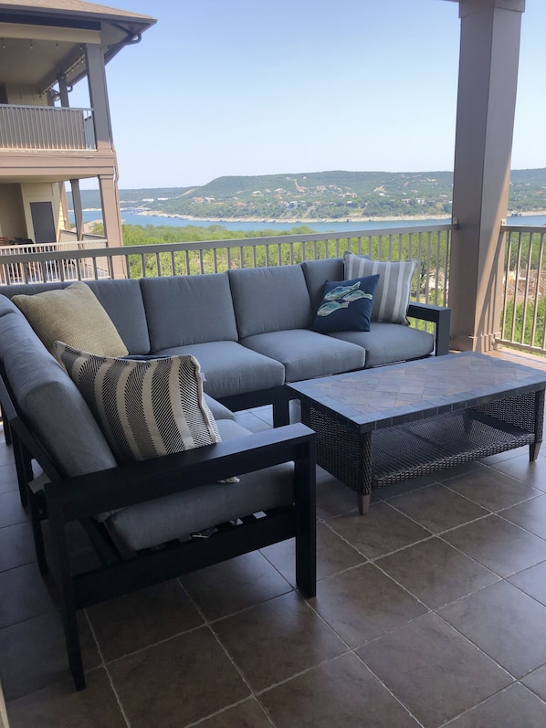 A balcony with a grey sofa and a black coffee table, overlooking a scenic view of a lake and hills.
