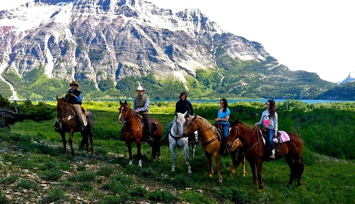 A group of people on horseback in a grassy field with a mountainous backdrop.