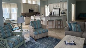 Un'ampia zona giorno con divano, poltrona e tavolino. Si vede una cucina con mobili bianchi e una zona pranzo con tavolo e sedie.