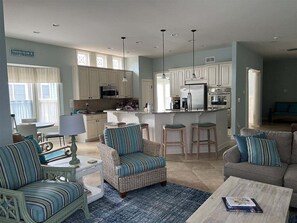 Un'ampia zona giorno con divano, poltrona e tavolino. Si vede una cucina con mobili bianchi e una zona pranzo con tavolo e sedie.