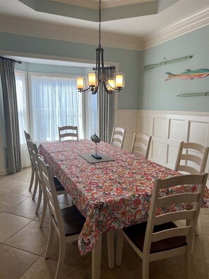 Zona pranzo con tavolo ricoperto da una tovaglia floreale, circondato da sedie di legno, un lampadario e una finestra con tende.