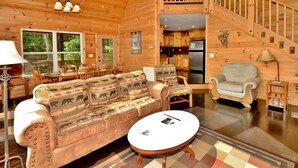 Un intérieur de chalet chaleureux, avec un revêtement mural en bois, un canapé agrémenté de coussins à motifs animaliers, une table basse ronde et un escalier menant à un niveau supérieur.