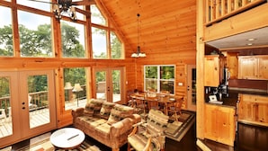 Une cabane en bois spacieuse, dotée de grandes fenêtres, d’un salon confortable avec un canapé et des fauteuils, d’une table à manger et d’un coin cuisine.