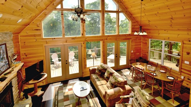 Un intérieur de chalet chaleureux avec un plafond haut, de grandes fenêtres, une cheminée, un canapé, une table à manger et un téléviseur.