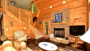 L’intérieur d’un chalet confortable en bois, avec une cheminée, un téléviseur, un coin salon confortable et un escalier.