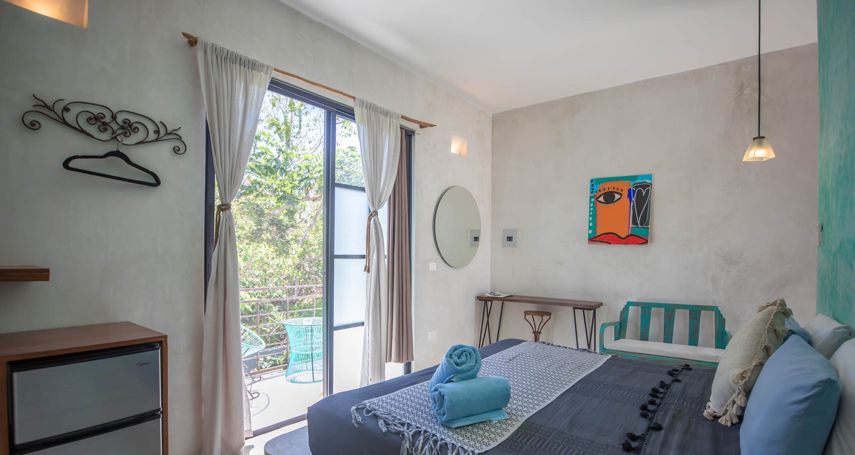 A bedroom with a bed, a desk, a chair, a hanging coat rack, and a window with curtains.