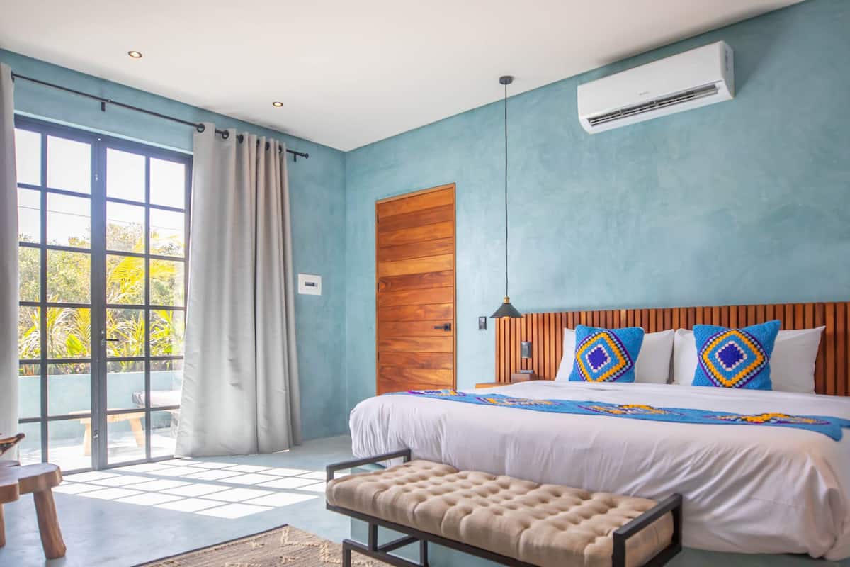 A bedroom with a large bed, a wooden headboard, a bench, and a view of greenery outside.