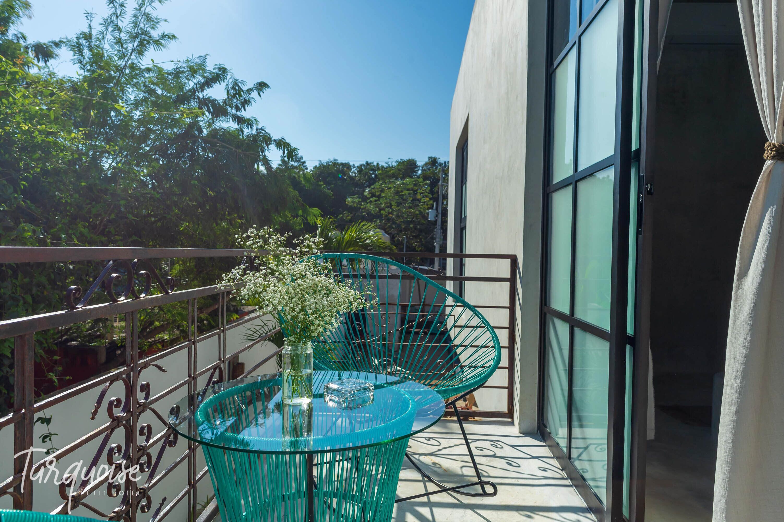 Un balcón con una mesa y sillas de vidrio, un florero con flores y vistas a árboles y un edificio.