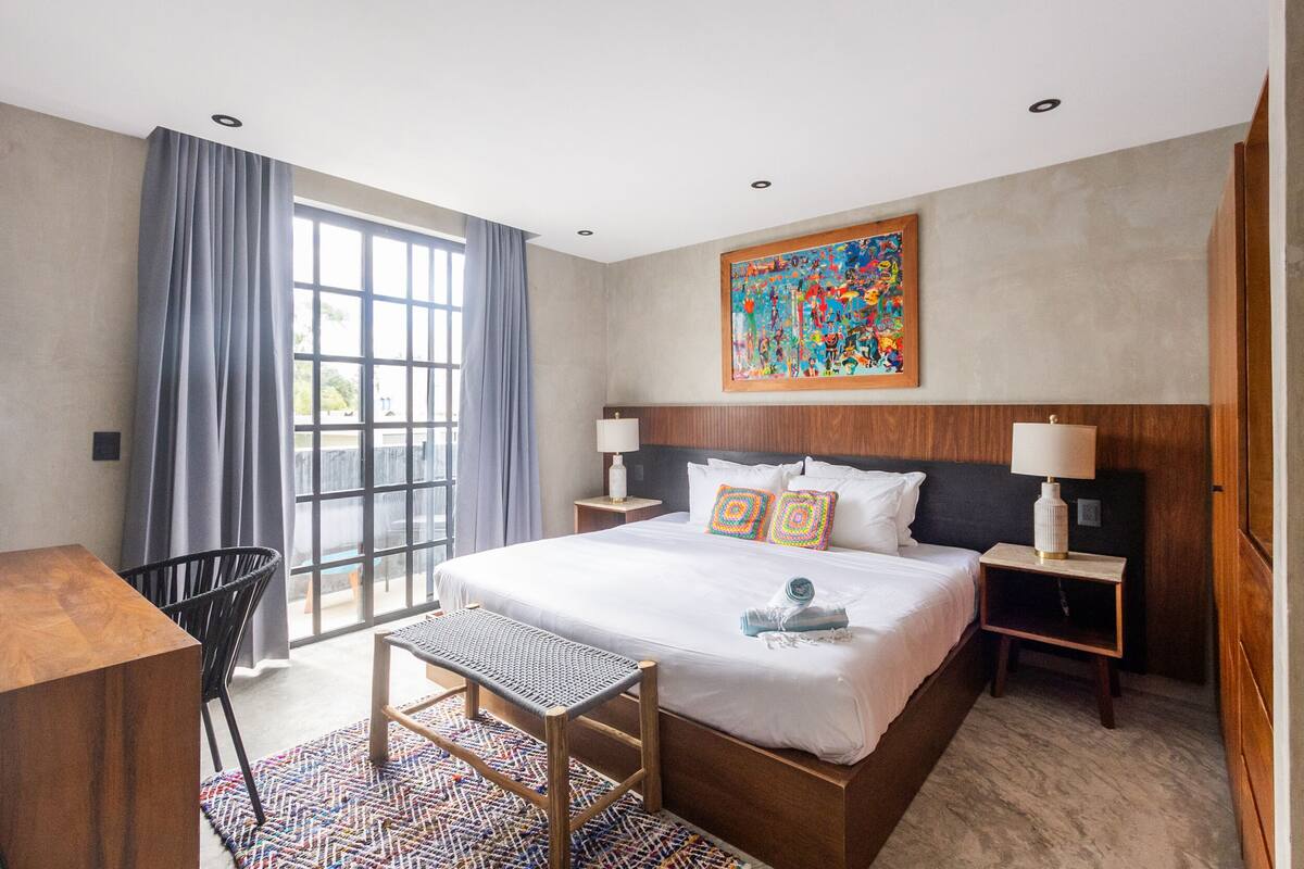 A hotel room with a large bed, two bedside tables, a desk, a chair, a window with curtains, and a colorful artwork on the wall.