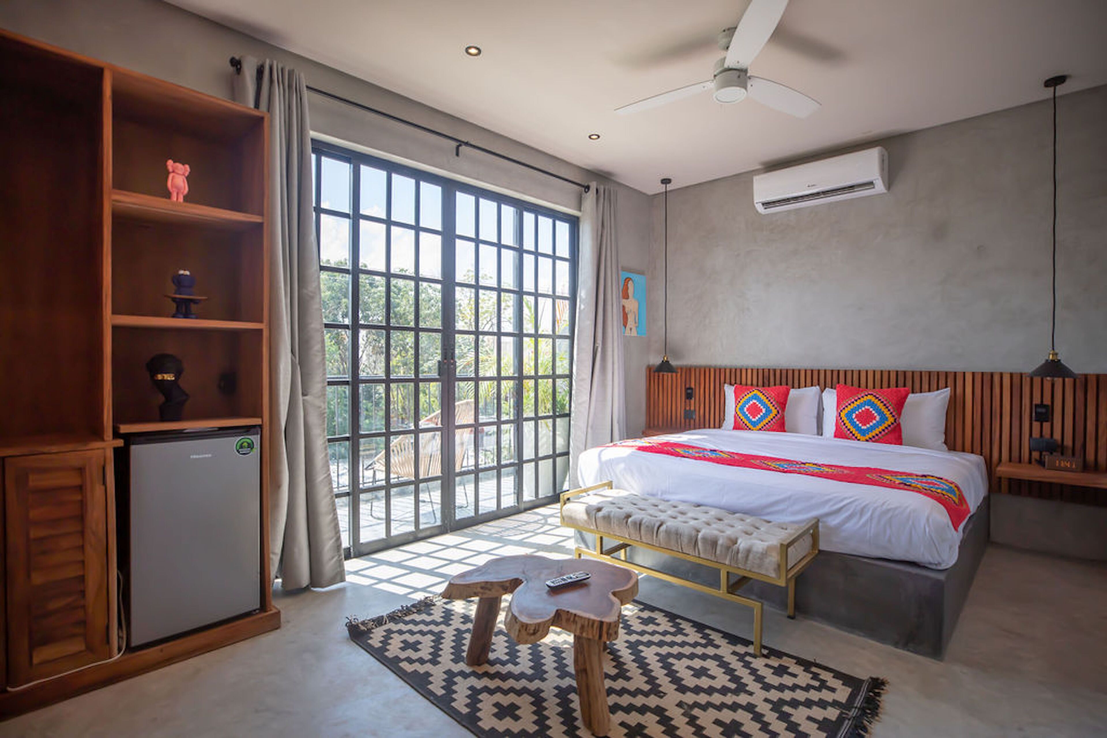 A modern bedroom with a large bed, a wooden wardrobe, a small refrigerator, and a view through a large window.