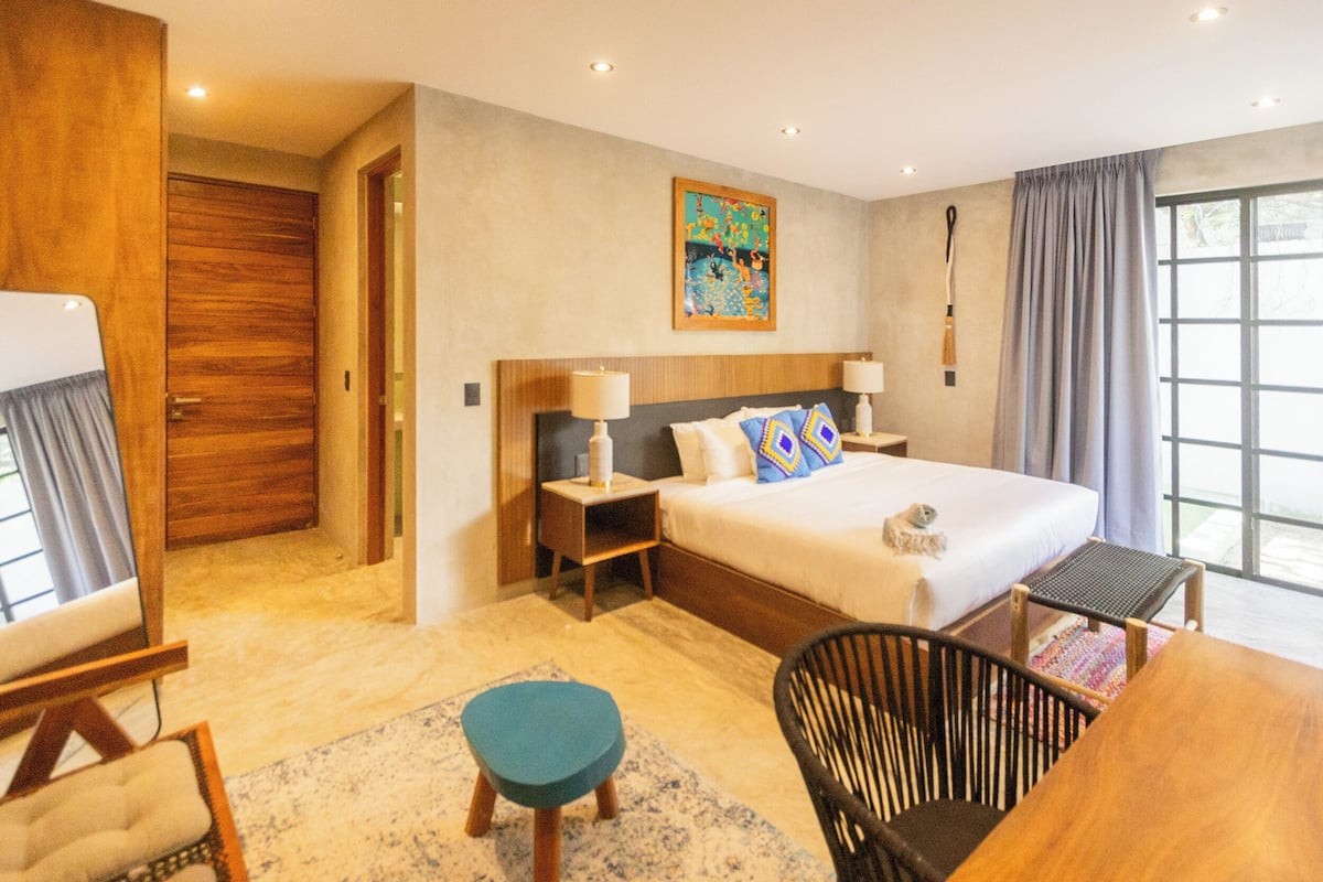 A modern hotel room with a large bed, two bedside tables, a desk, a chair, and a window with curtains.