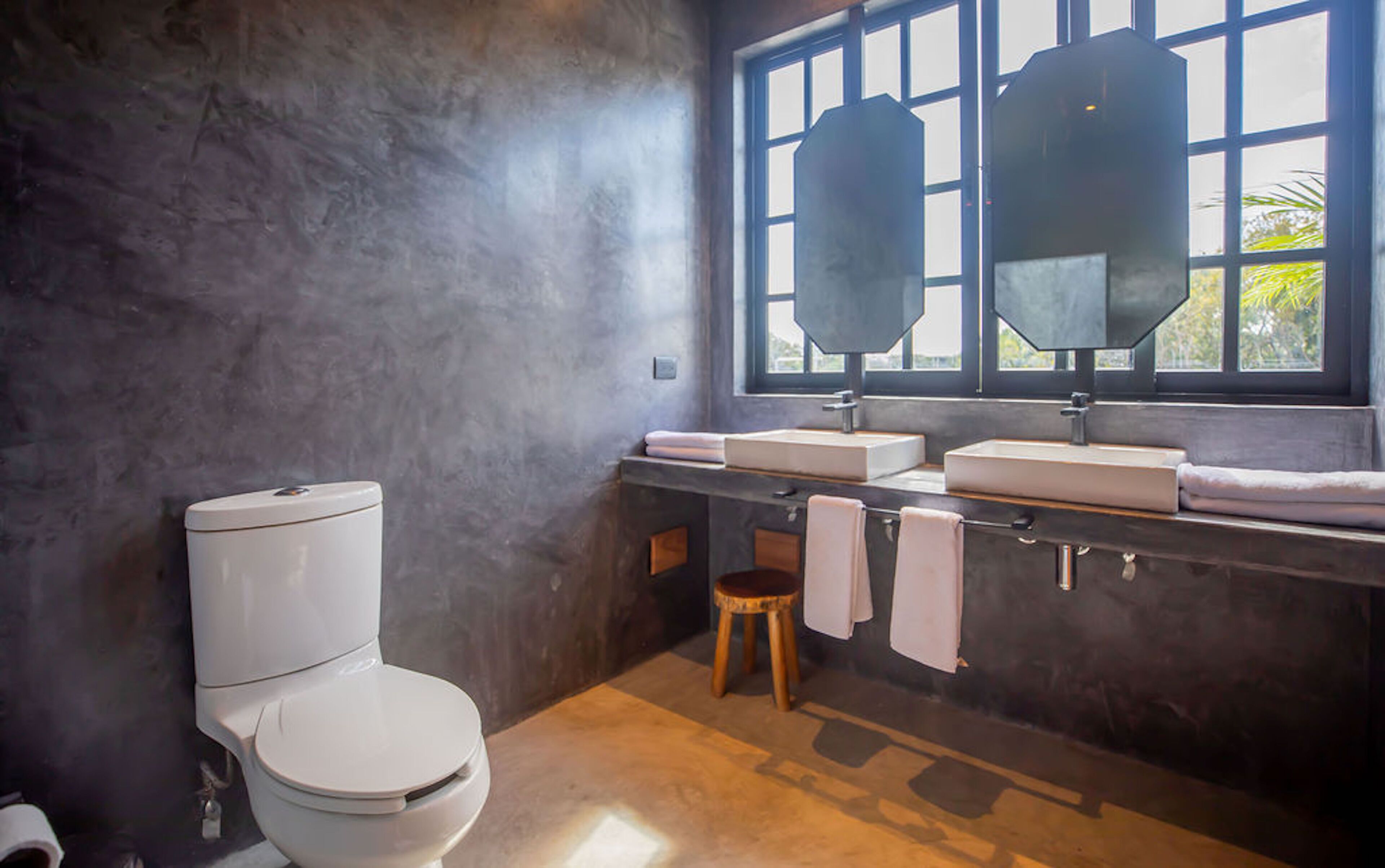 Un baño con inodoro, dos lavamanos, ventana y toallero.