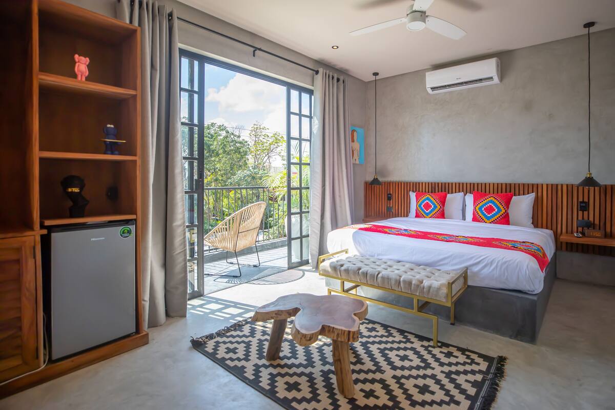 A modern bedroom with a large bed, a small desk, a ceiling fan, and a view of greenery outside.
