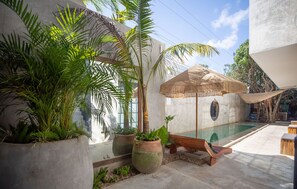 Zona de piscina con una tumbona, plantas en macetas grandes y un parasol de paja.