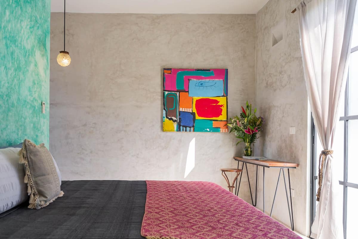 A modern bedroom with a bed, pillows, a colorful abstract painting, a vase with flowers, and a view of the outdoors through a window.