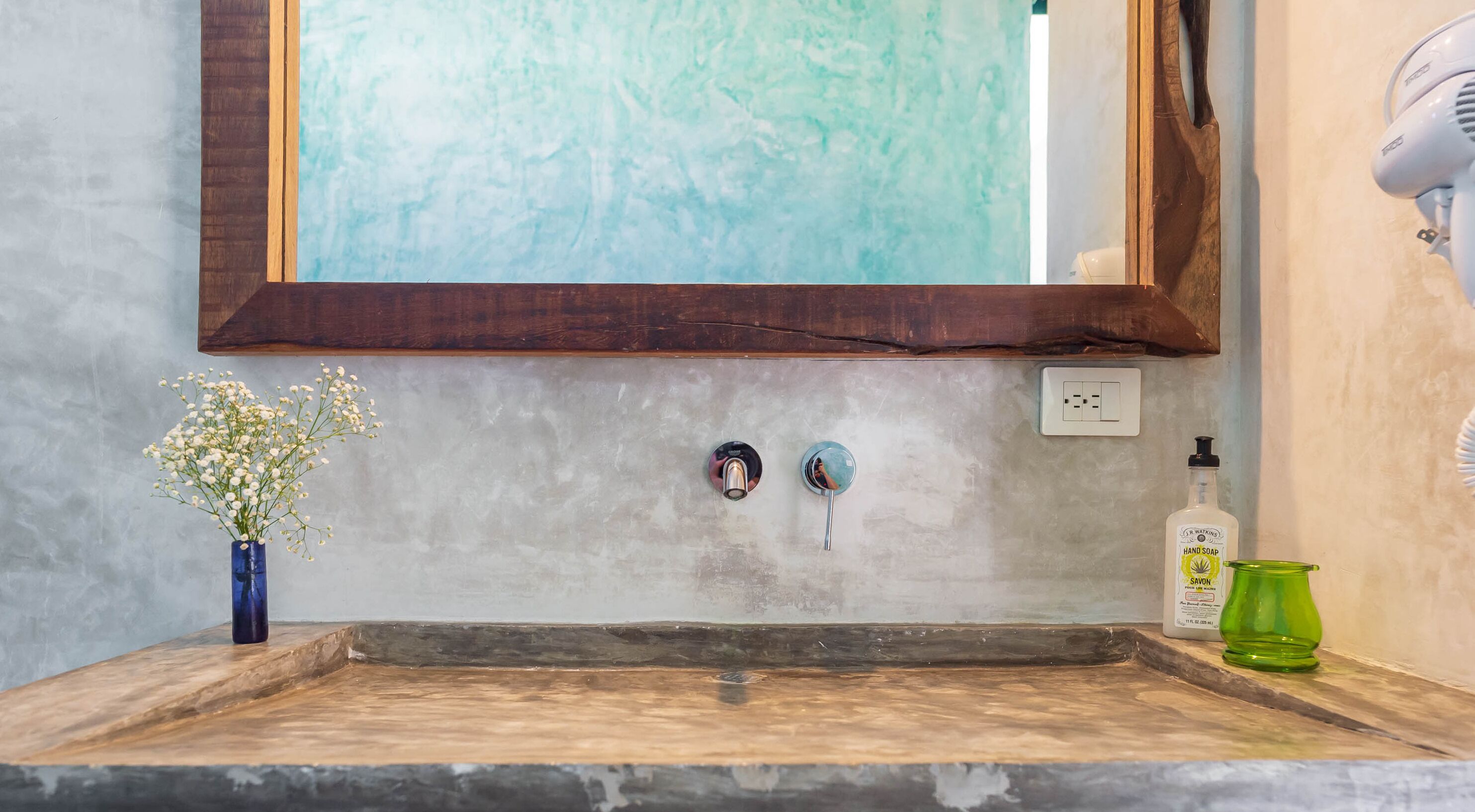 A bathroom with a wooden-framed mirror, a sink with two faucets, a vase with flowers, a green glass bottle, and a hand soap dispenser.
