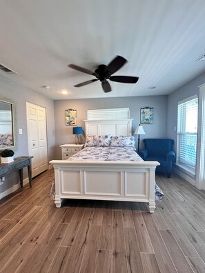 A bedroom with a large bed, a ceiling fan, a blue armchair, and a window with blinds.