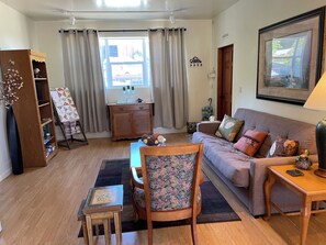Ein gemütliches Wohnzimmer mit einer Couch, einem Holztisch, einem Beistelltisch, einem Bücherregal und einem Fenster mit Vorhängen.