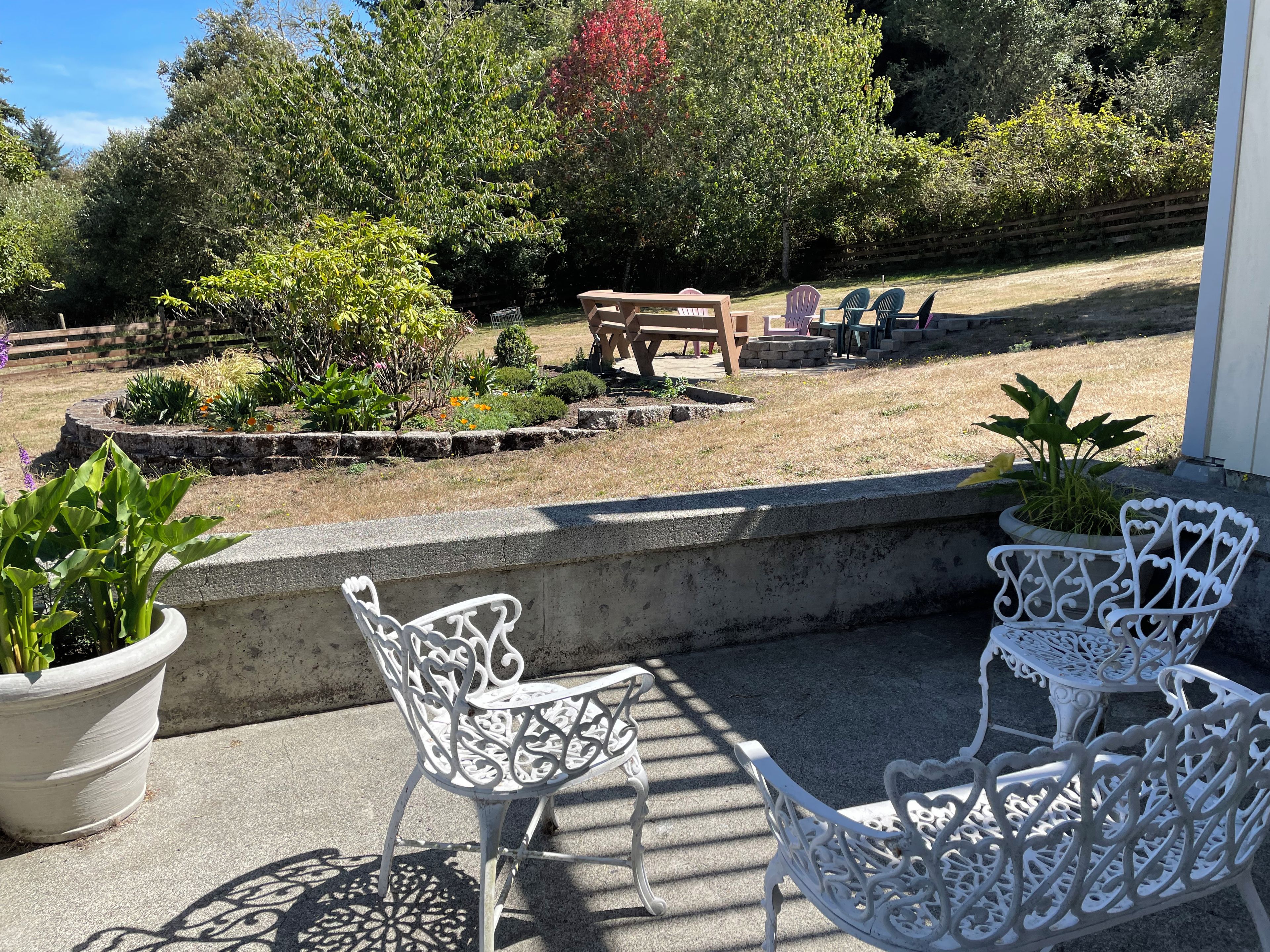 A patio with white metal chairs and a table, surrounded by a garden with various plants and a wooden bench in the background.