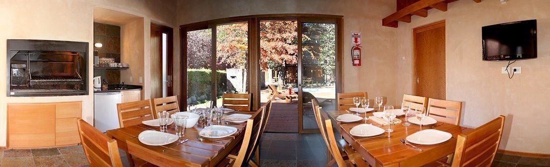 Un comedor con una mesa larga dispuesta para cuatro, una parrilla empotrada y un televisor en la pared.