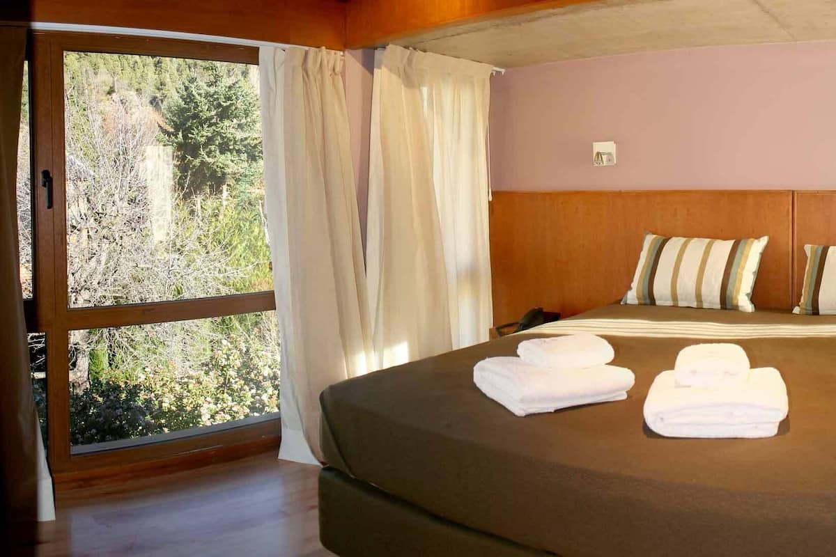 A bedroom with a large bed, two pillows, a wooden nightstand, and a window with curtains.