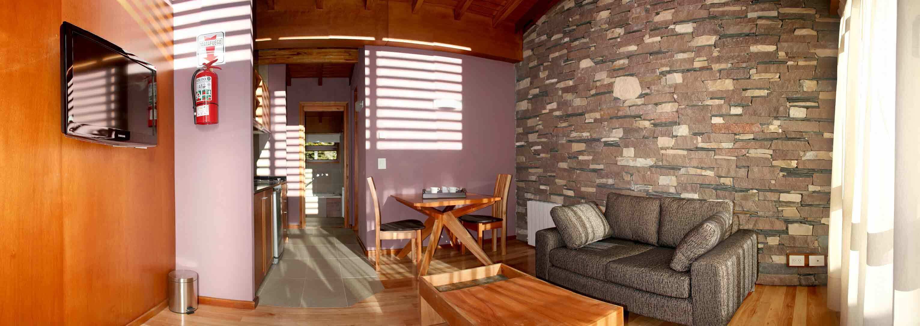 A cozy living area with a stone wall, a sofa, a coffee table, and a dining table with chairs. A fire extinguisher is mounted on the wall.