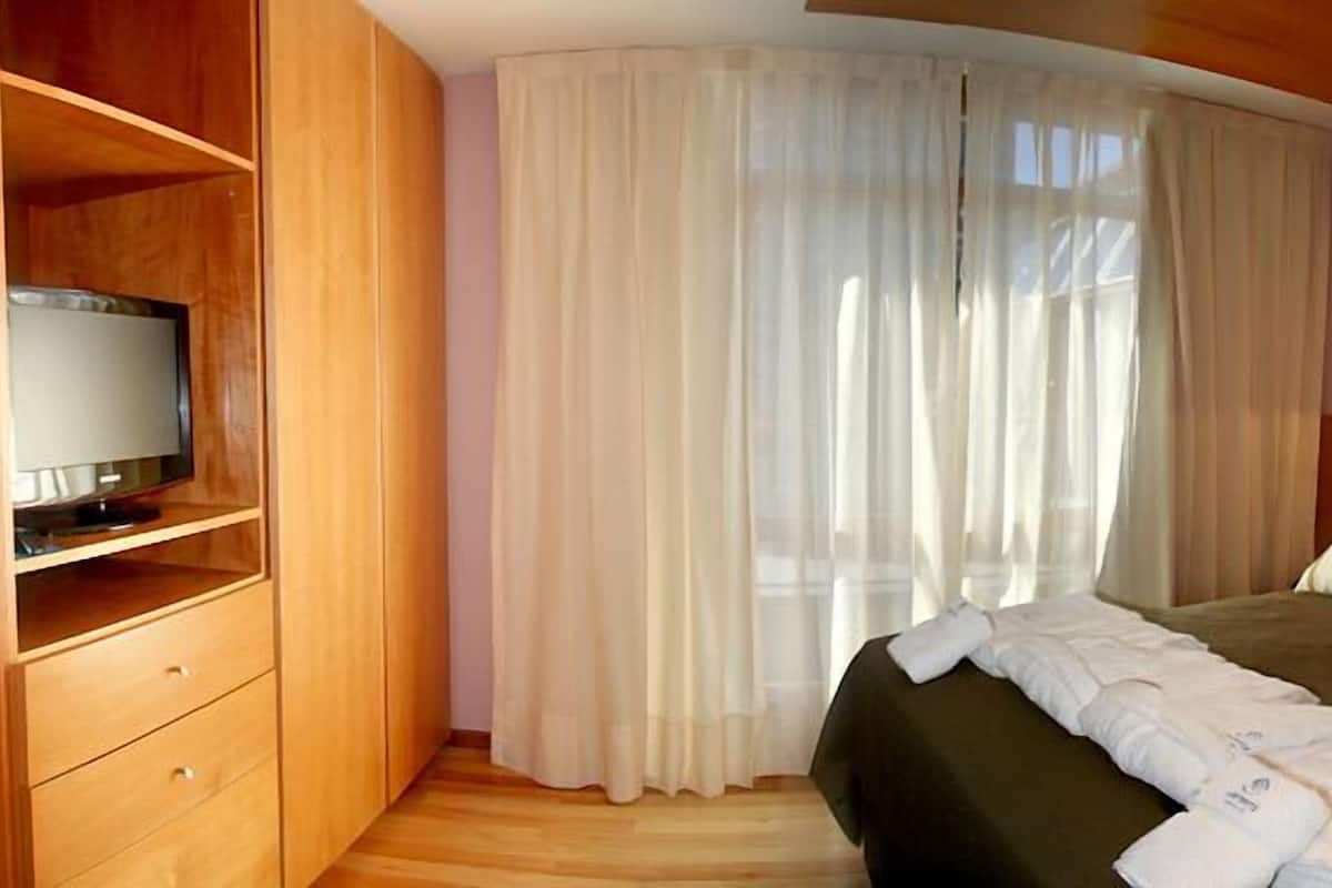 A hotel room with a bed, a TV on a wooden shelf, a nightstand, and a window with sheer curtains.