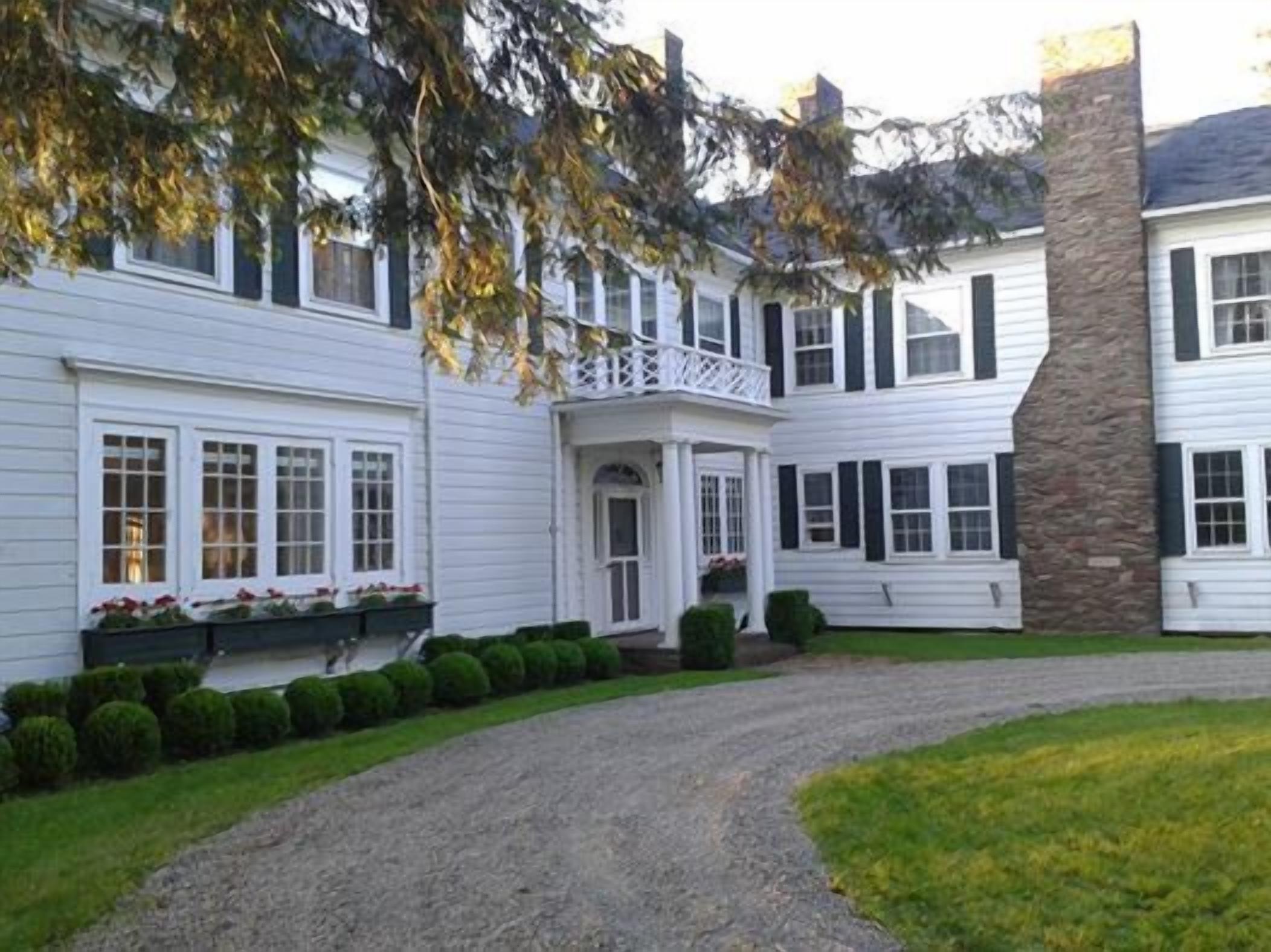A two-story white house with a stone chimney, a covered porch, and a gravel driveway.