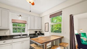Una cocina con armarios blancos, una mesa de madera y un banco. Se ve vegetación a través de una ventana.