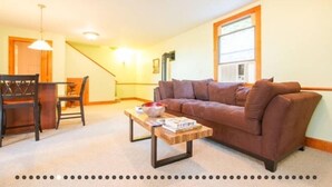 A living room with a brown sofa, a wooden coffee table, a dining table with chairs, and a staircase in the background.