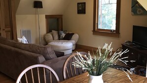 Un comedor con una mesa de madera, sillas y un jarrón de flores. Se observa un sofá y un televisor al fondo.