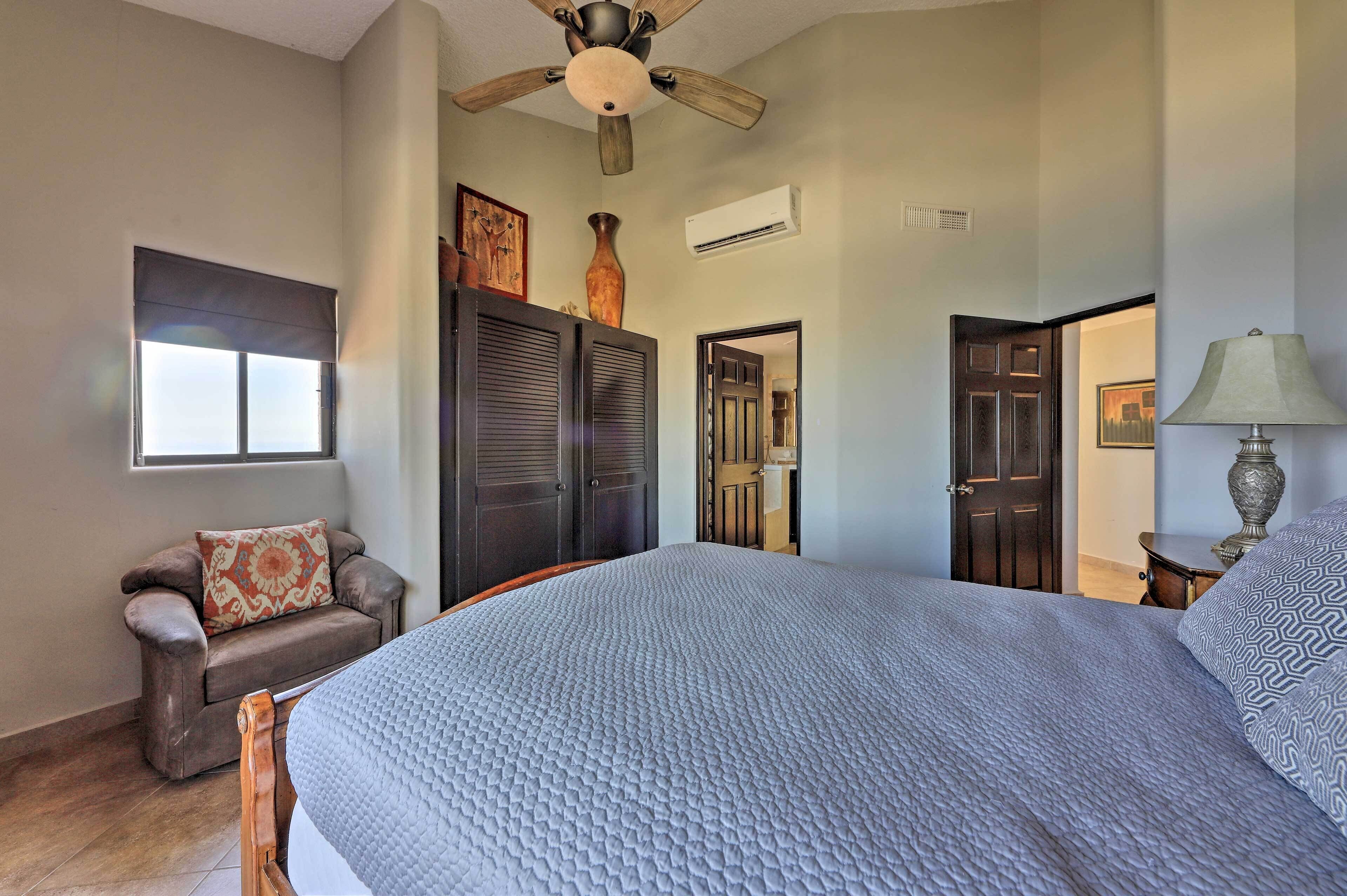 A bedroom with a large bed, a ceiling fan, a window with blinds, a dark wooden wardrobe, and a nightstand with a lamp.