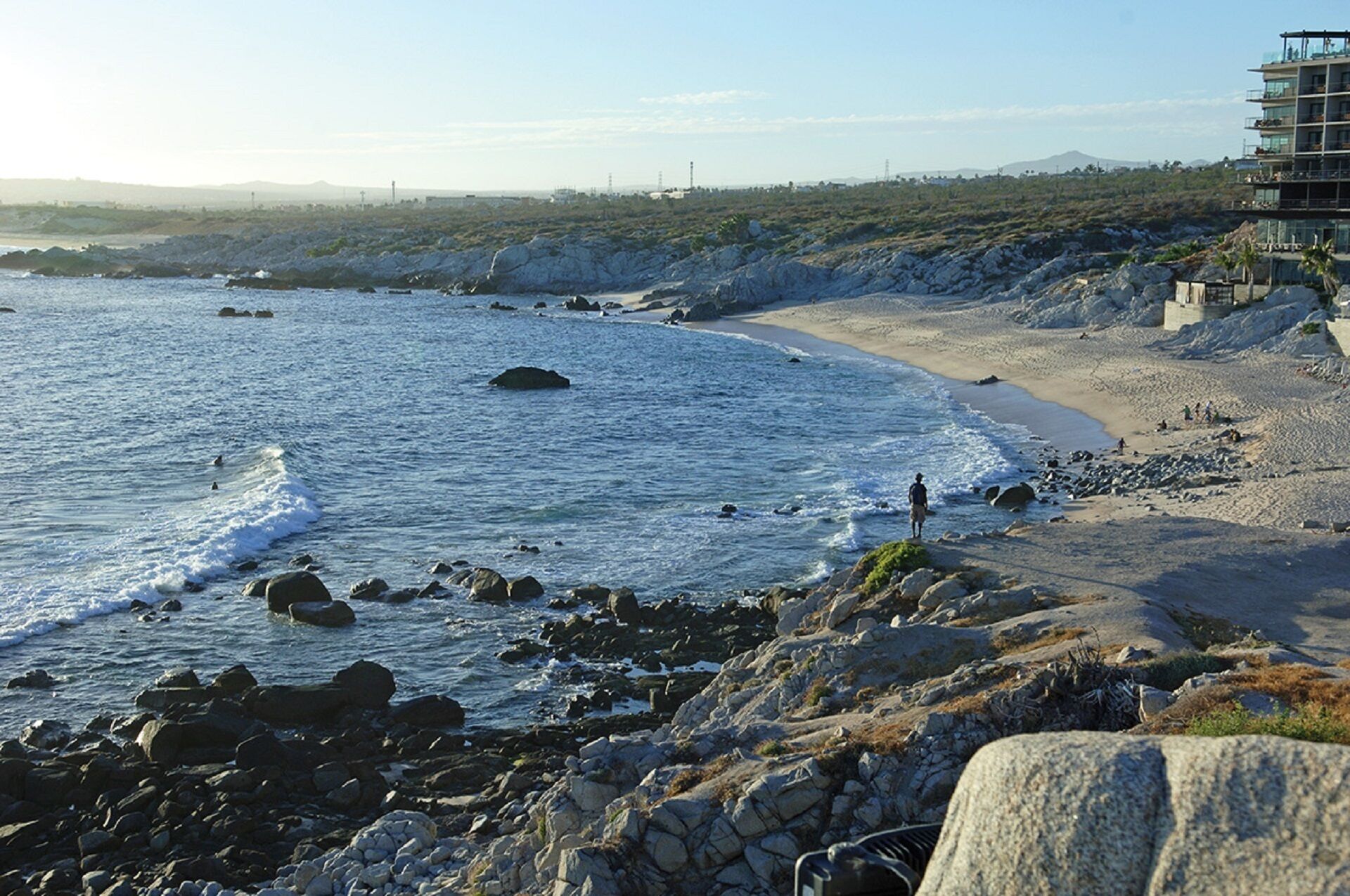 En strand med klippefyldt terræn og en bygning med balkoner med udsigt over havet.