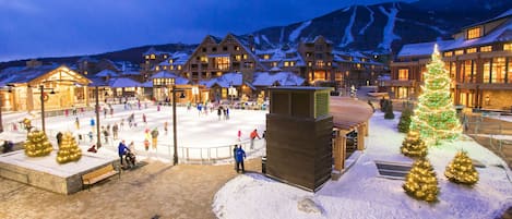 Un espacio exterior cubierto de nieve con un gran árbol de Navidad, edificios de madera y una pista de esquí al fondo.