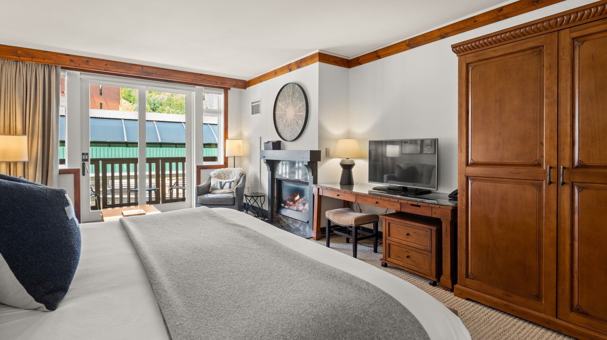A hotel room with a large bed, a desk with a TV, a fireplace, and a view of the outside balcony.