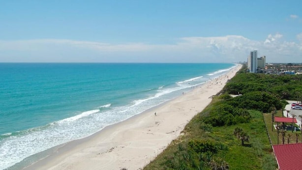 Ein Strand mit sandigem Ufer, einem klaren blauen Ozean und einigen Gebäuden im Hintergrund.