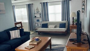 Ein Wohnzimmer mit einem dunkelblauen Sofa, einem Holztisch und einem weißen Sofa. Ein Fernseher steht auf einem Holzgestell, eine Lampe und ein Regal mit Dekorationen sind ebenfalls vorhanden.