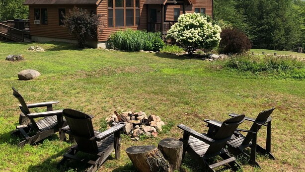 A wooden cabin with a large chimney, a deck, and a fire pit area with chairs. - Beautiful, family-friendly home in private wilderness setting (Wilmington)