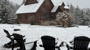 A snow-covered landscape with a large brick house, three Adirondack chairs, and a snow-covered tree. - Beautiful, family-friendly home in private wilderness setting (Wilmington)