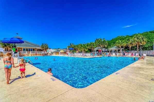 A swimming pool area with lounge chairs and umbrellas, surrounded by palm trees and a building with a sloped roof.