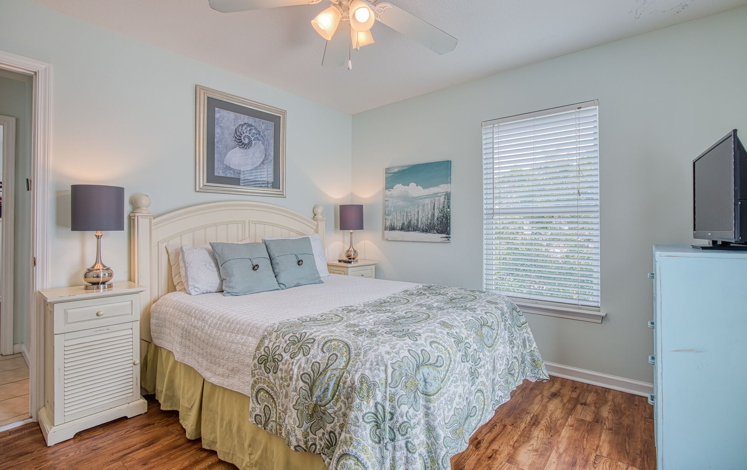 A bedroom with a bed, two bedside tables, a ceiling fan, a window with blinds, and a TV on a cabinet.