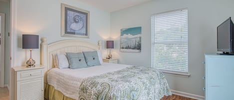 A bedroom with a bed, two bedside tables, a ceiling fan, a window with blinds, and a TV on a cabinet.