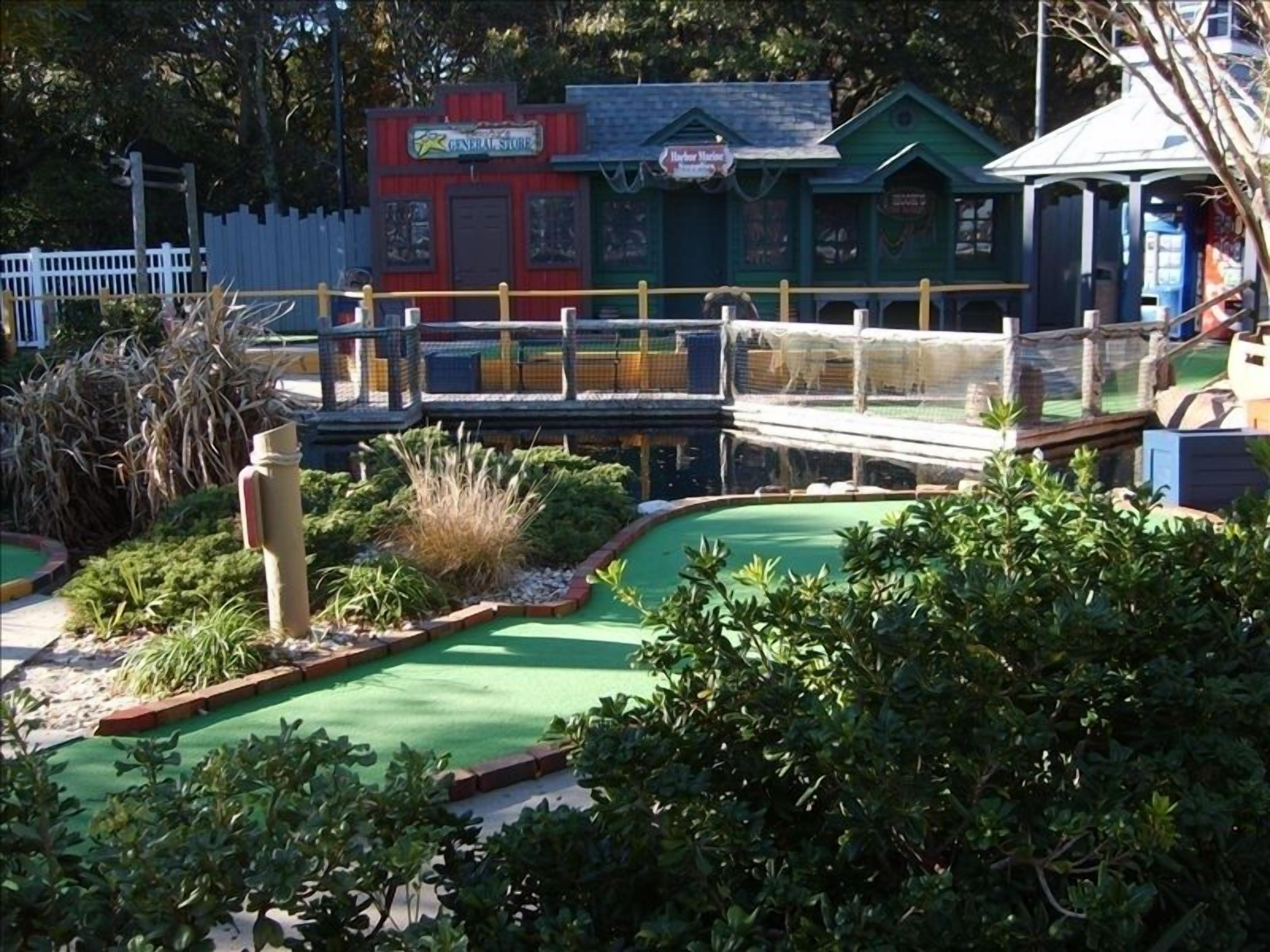 Un campo da mini golf con una zona verde per il gioco, un edificio rosso con la scritta 'General Store' e un edificio blu con il cartello 'Harbor Marine Services'.
