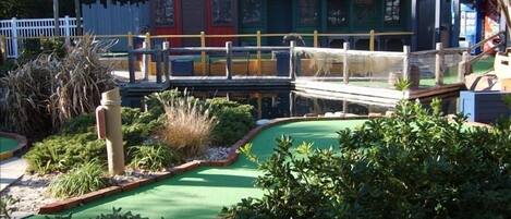 Un campo da mini golf con una zona verde per il gioco, un edificio rosso con la scritta 'General Store' e un edificio blu con il cartello 'Harbor Marine Services'.