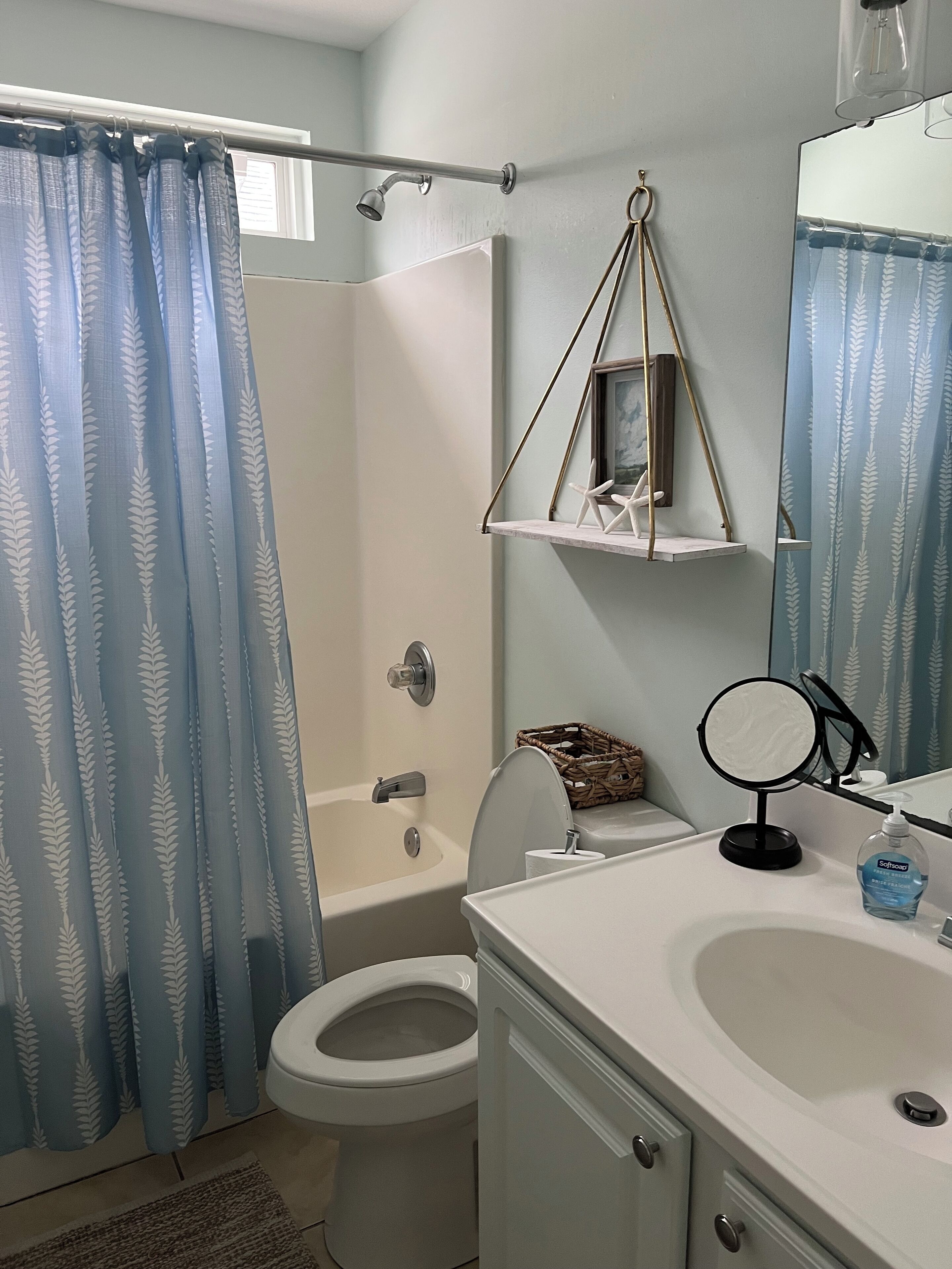 A bathroom with a shower, toilet, and sink. There is a hanging shelf with a framed picture and a small mirror.