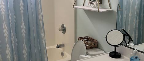 A bathroom with a shower, toilet, and sink. There is a hanging shelf with a framed picture and a small mirror.