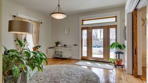 A spacious living room with a large window, a wooden floor, a round rug, and several potted plants.