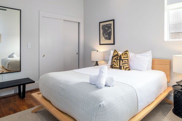 A neatly made bed with white linens and rolled towels, a bedside table with a lamp, a wall-mounted picture, and a door.